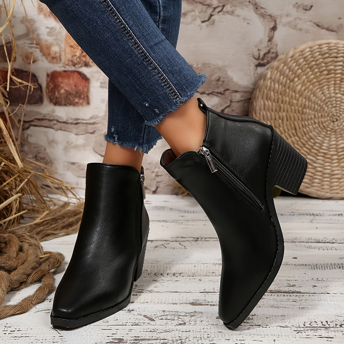 A pair of women's black faux-leather ankle boots with a side zipper and block heel, shown on a model wearing blue jeans in a rustic styled setting.