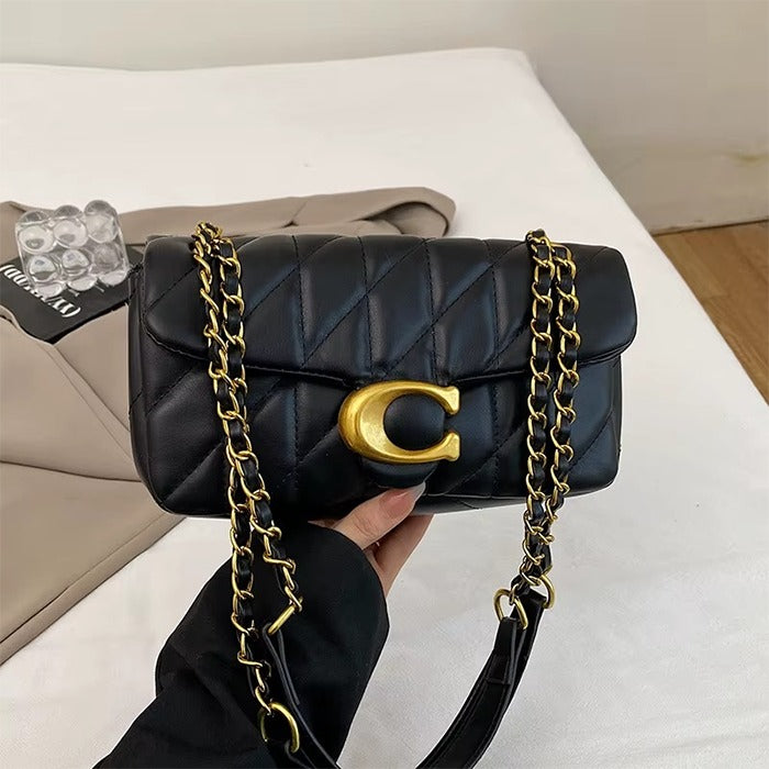 A black quilted leather handbag with a gold-tone C-shaped hardware clasp and a gold chain strap, held by a hand against a white background with a beige blazer and clear acrylic blocks in the background.