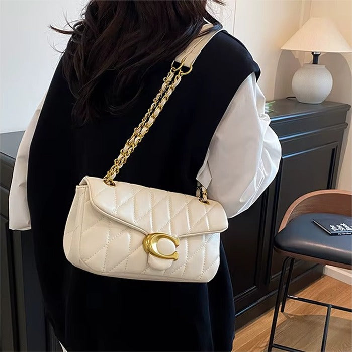 A woman wearing a black vest over a white long-sleeve shirt carries a cream quilted shoulder bag with a gold chain strap and a gold C-shaped logo clasp. The setting is a modern indoor room with a dark cabinet, a lamp, and a chair.