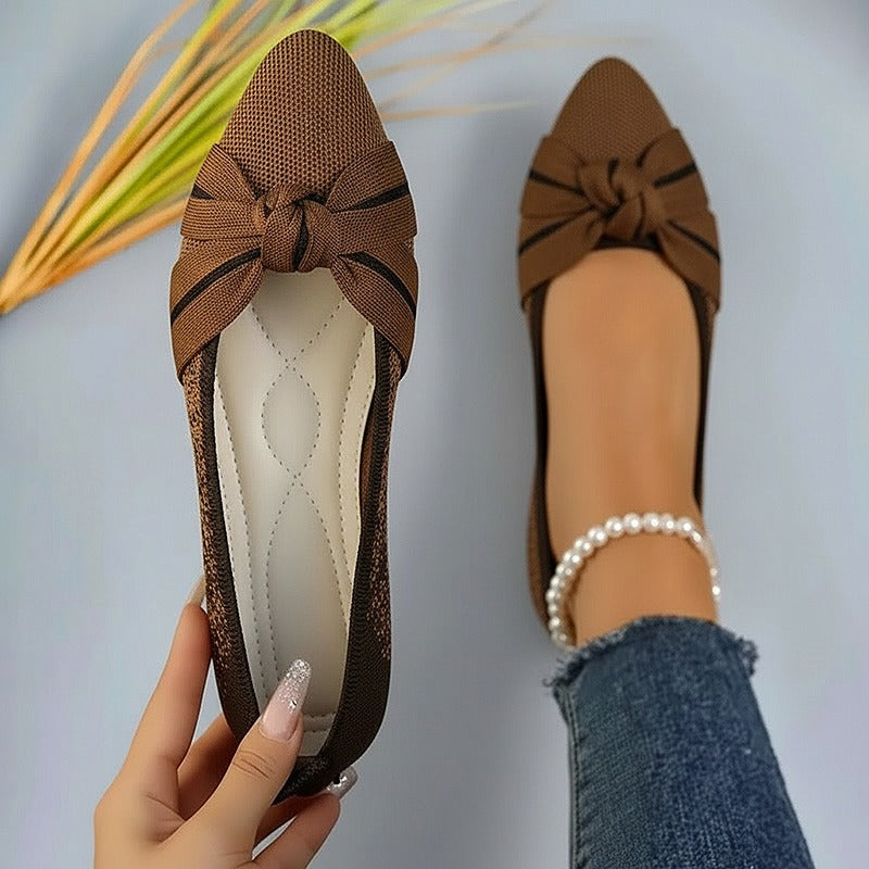 Brown flat shoes with a bow held by a hand against a neutral background