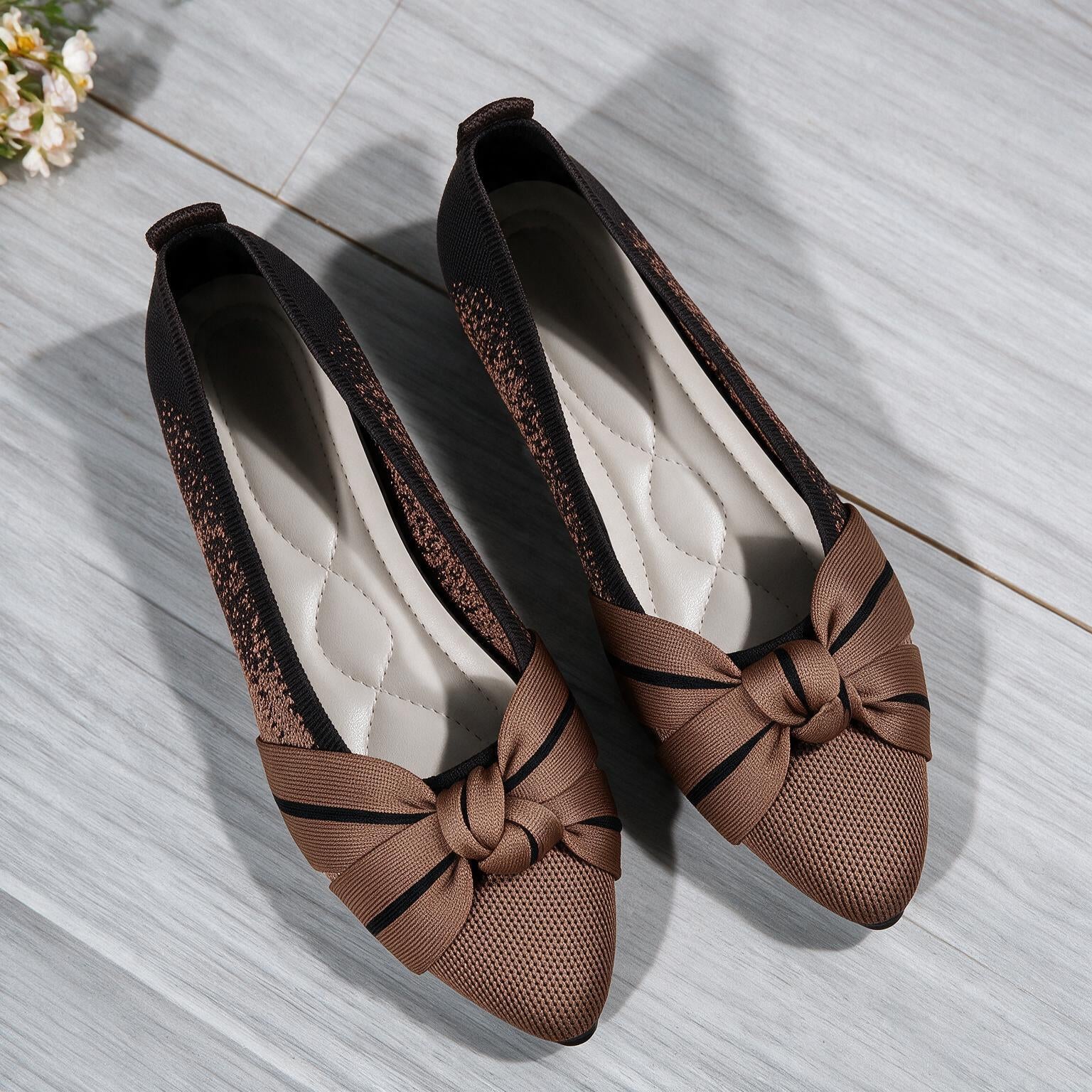 A pair of women's slip-on flats with pointed toes, featuring a brown mesh upper, a wide brown satin bow with black stripes, and a black glittery textured side panel, displayed on a light gray wood floor.