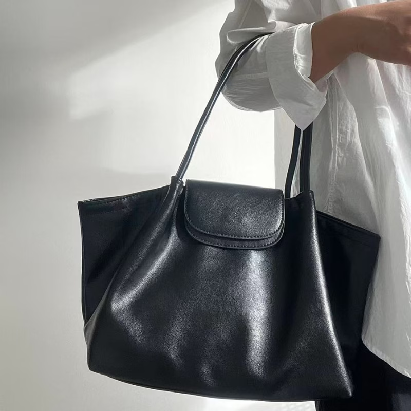 A black leather tote bag with a top flap and long shoulder straps, held by a person wearing a white shirt against a plain light background.