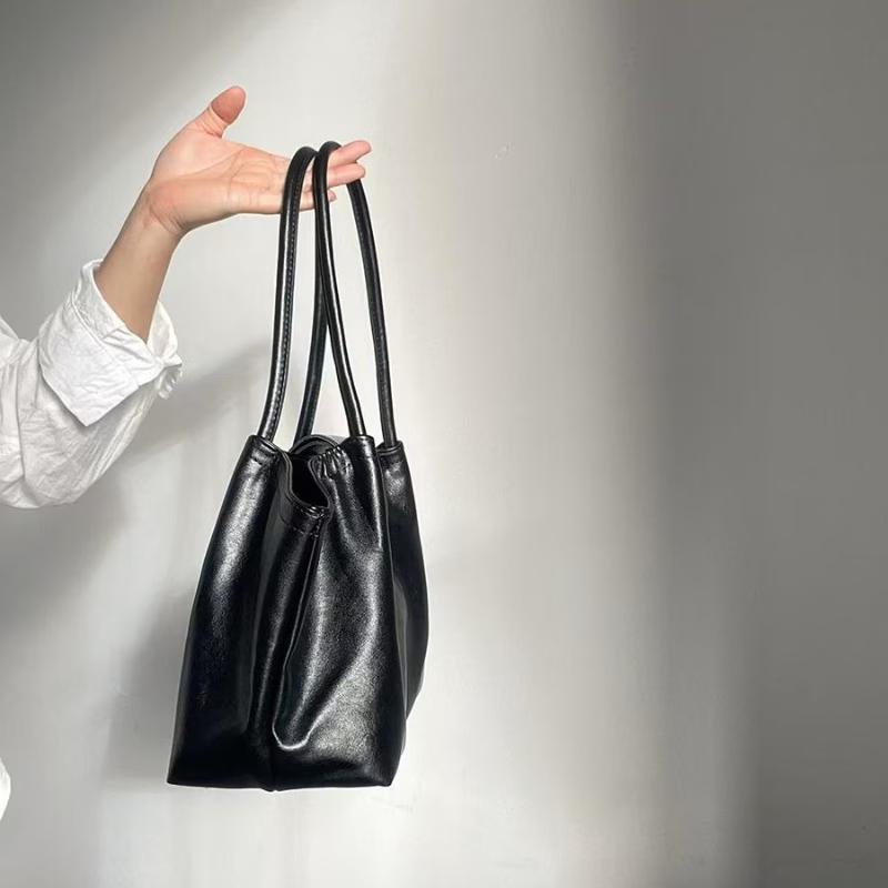 A black leather bucket bag with double shoulder straps, held by a person's hand against a plain light gray wall.
