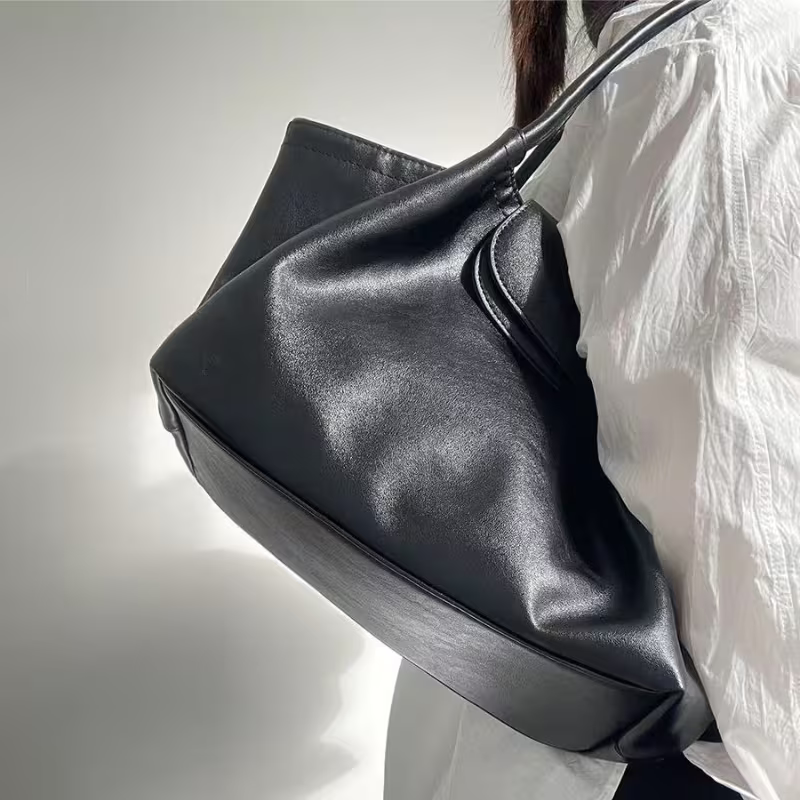 A large black leather slouchy shoulder bag being worn by a person wearing a white wrinkled blouse, shown against a plain light gray background.