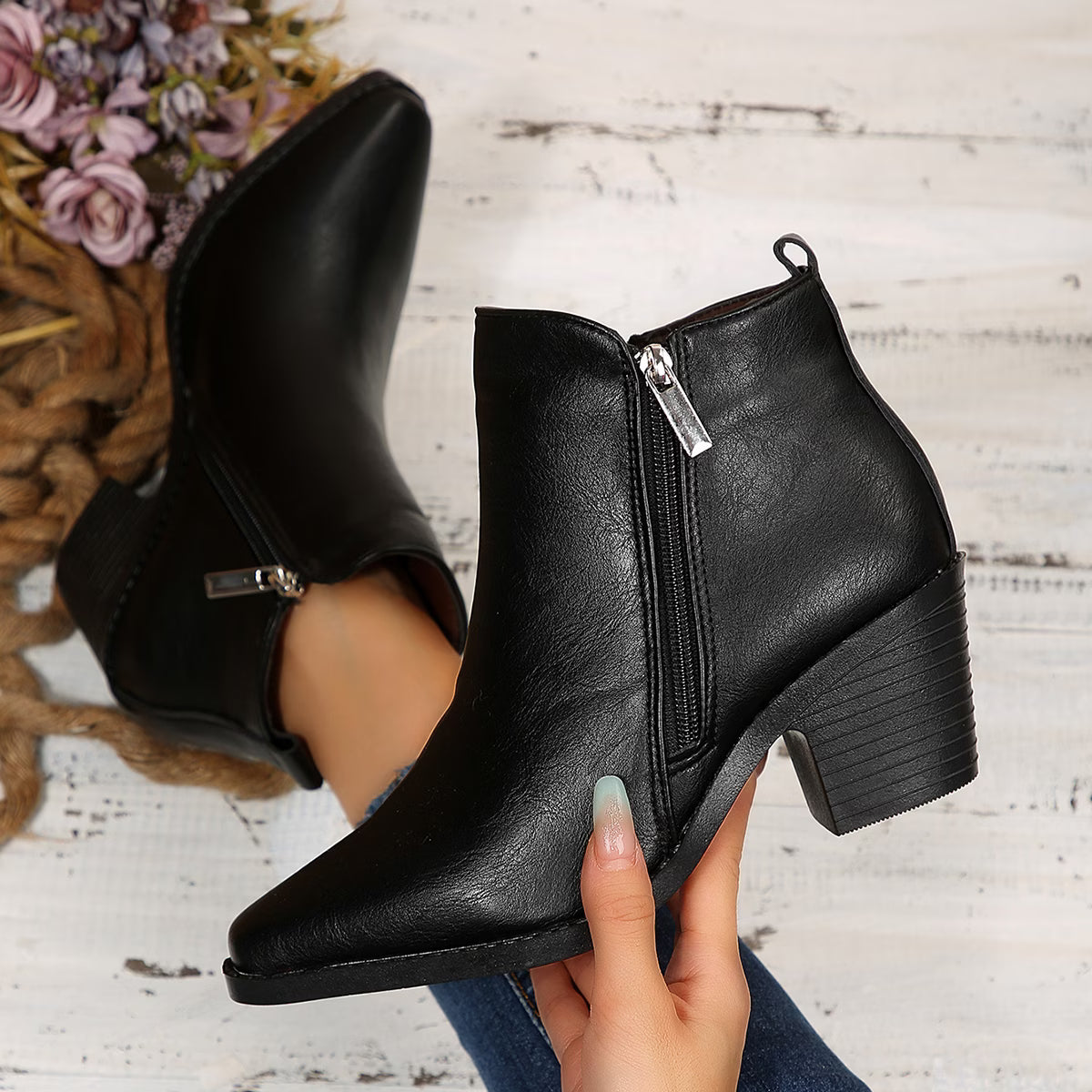 Black leather ankle boots with side zippers and block heels, shown on a rustic white wood background with a hand holding one boot and another boot resting on a surface nearby.