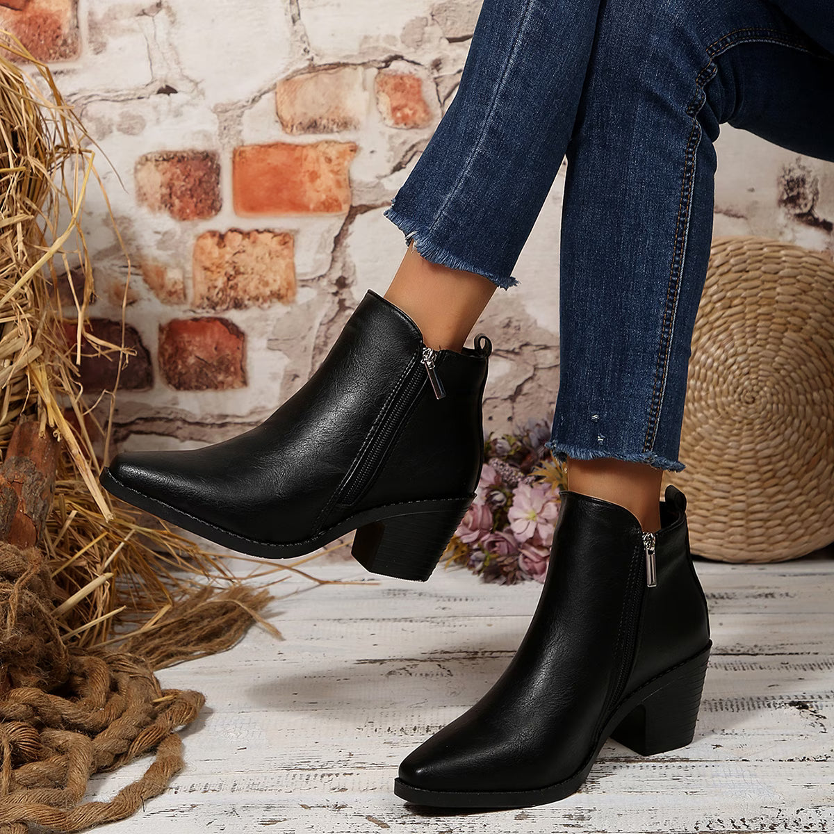 A pair of women's black faux-leather ankle boots with a low block heel and side zipper, shown on a model wearing blue jeans in a rustic setting with a brick wall, straw rope, and woven basket.