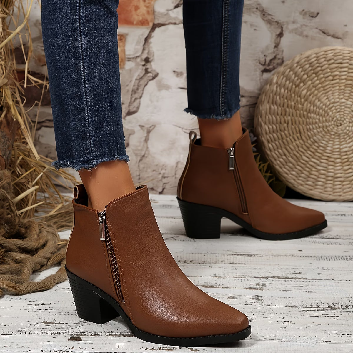 A pair of women's brown faux-leather ankle boots with a side zipper and low block heel, shown on a model wearing blue jeans in a rustic styled setting.