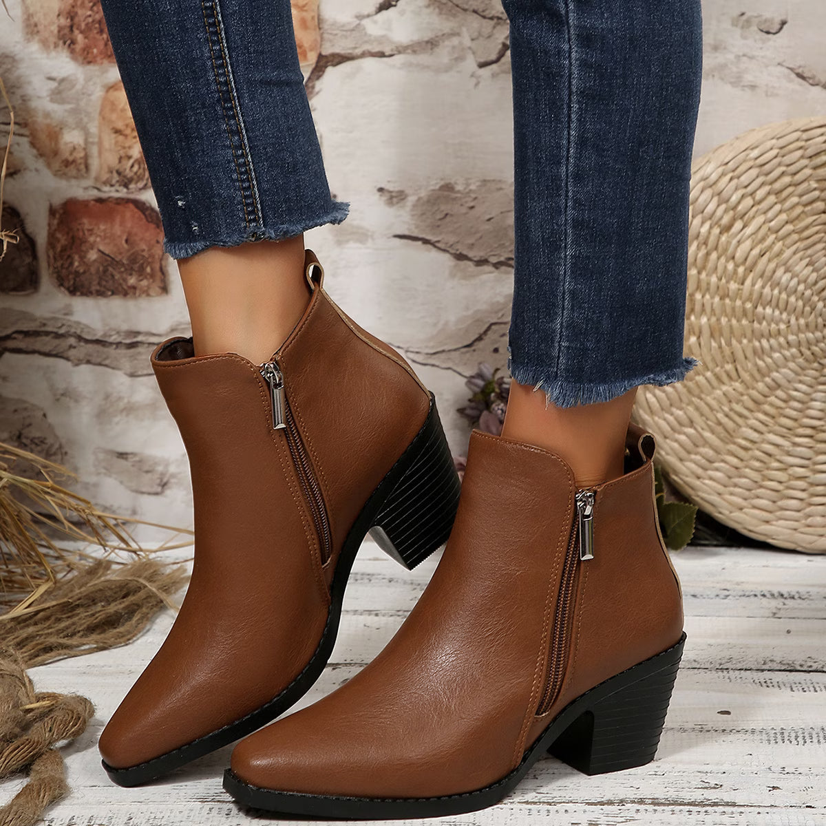 A pair of women's brown faux-leather ankle boots with a side zipper, black block heel, and pointed toe, shown on a model wearing blue jeans against a rustic brick and wood background.