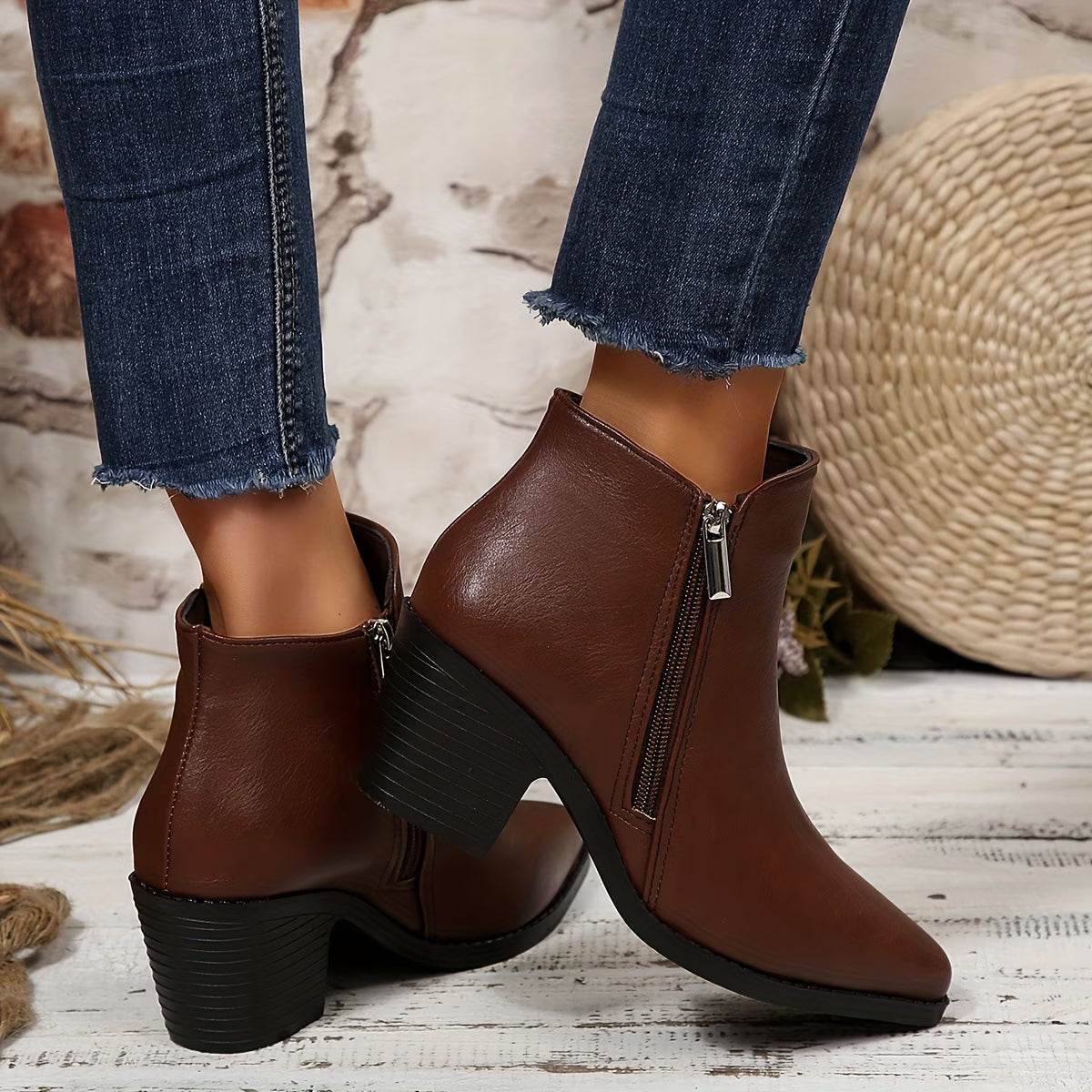 A pair of women's brown faux-leather ankle boots with black block heels and side zippers, shown on a model wearing blue jeans in a rustic styled setting.