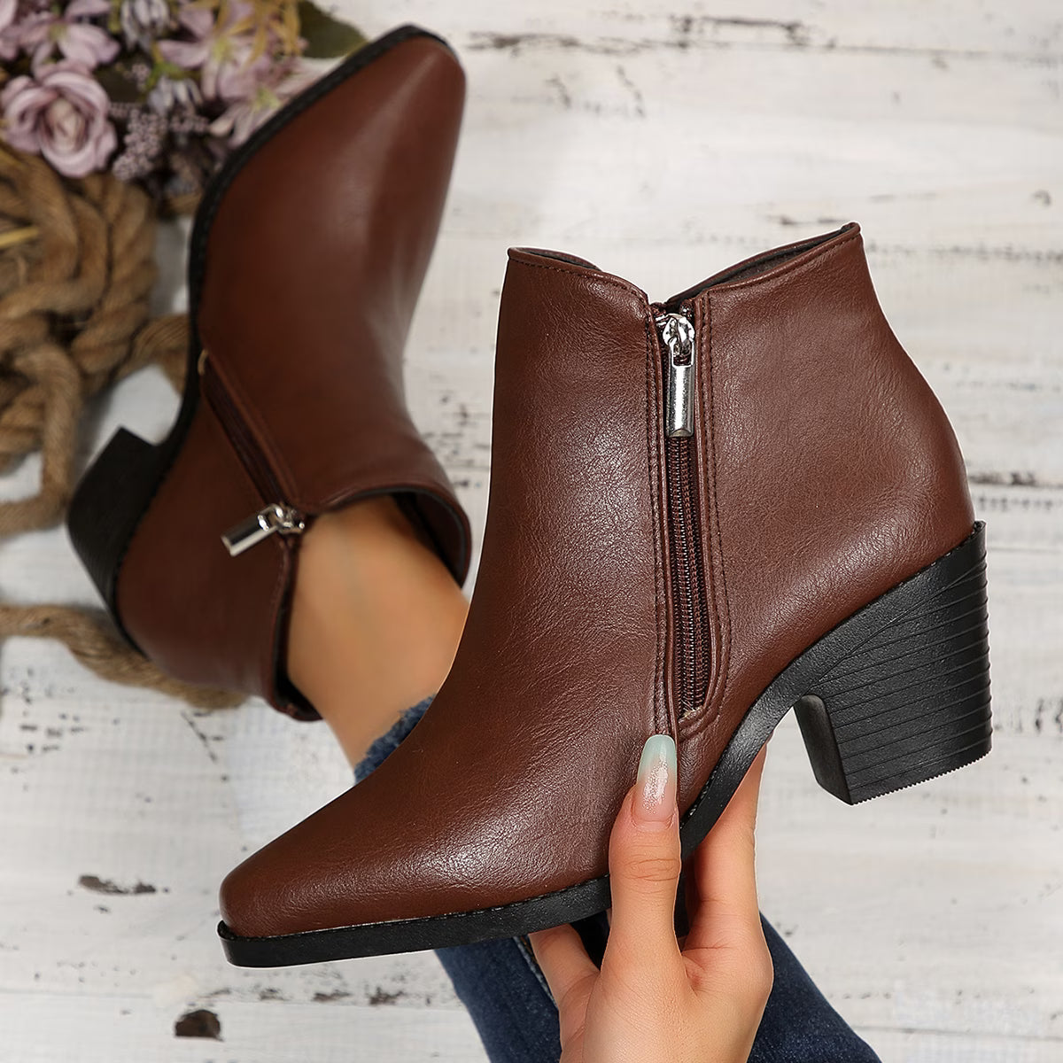 A pair of brown faux-leather ankle boots with side zippers and block heels, shown on a person's foot and held by a hand against a rustic white wood background.