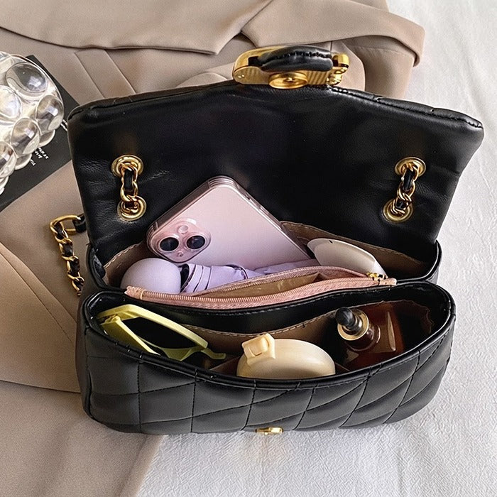 A black quilted leather handbag shown open, filled with various accessories including a pink phone, earbuds case, sunglasses, and cosmetics. The bag features gold-tone hardware and is set against a neutral beige background.