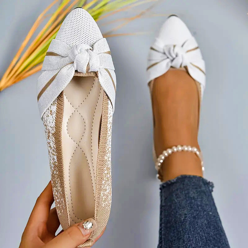 White pointed-toe shoes with a bow detail worn by a person, on a neutral background.