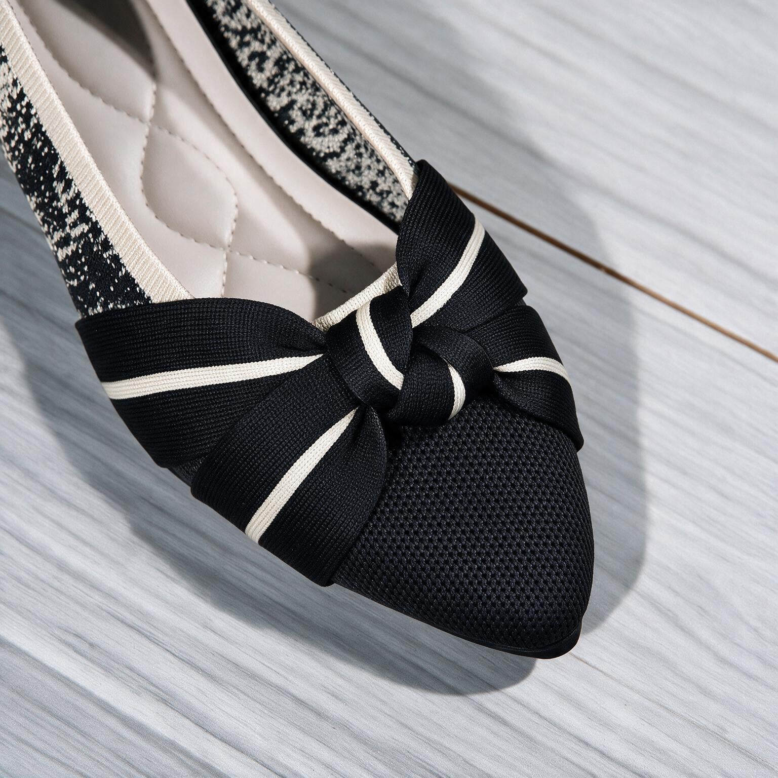 A close-up of a black slip-on shoe with a large bow featuring white stripes, a black mesh toe cap, and a glittery black-and-white side panel, shown on a light gray wood-textured surface.