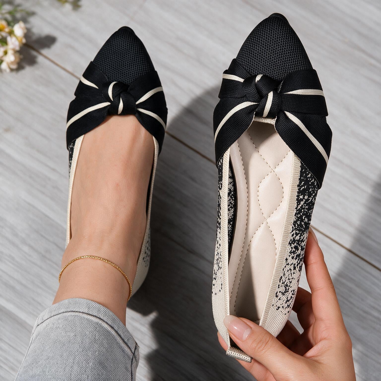 Black and cream flat shoes with a bow detail on the toe, shown on a model's foot and held in a hand against a gray wood floor background.