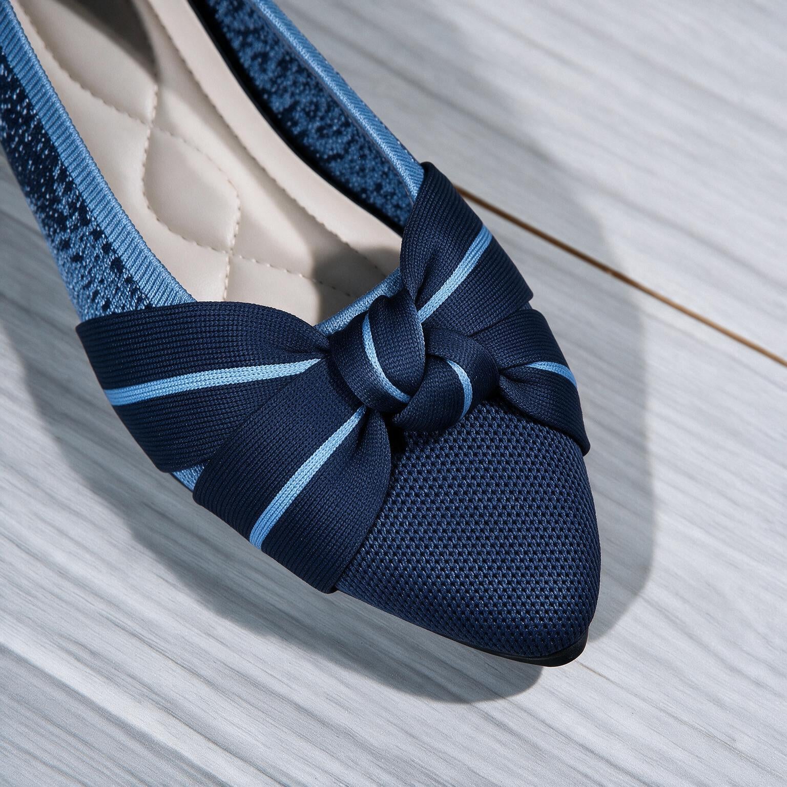 A close-up of a navy blue flat shoe with a textured mesh toe, a large matching fabric bow with light blue stripes, and a quilted cream insole, set on a light gray wood floor.