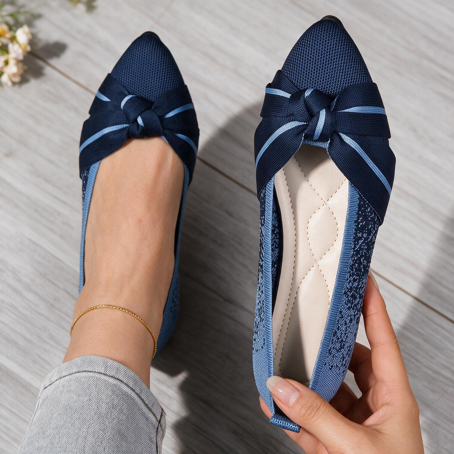 A pair of navy blue flat shoes with a knotted bow detail on the toe, shown on a model's foot and held in a hand against a light gray wood floor background.