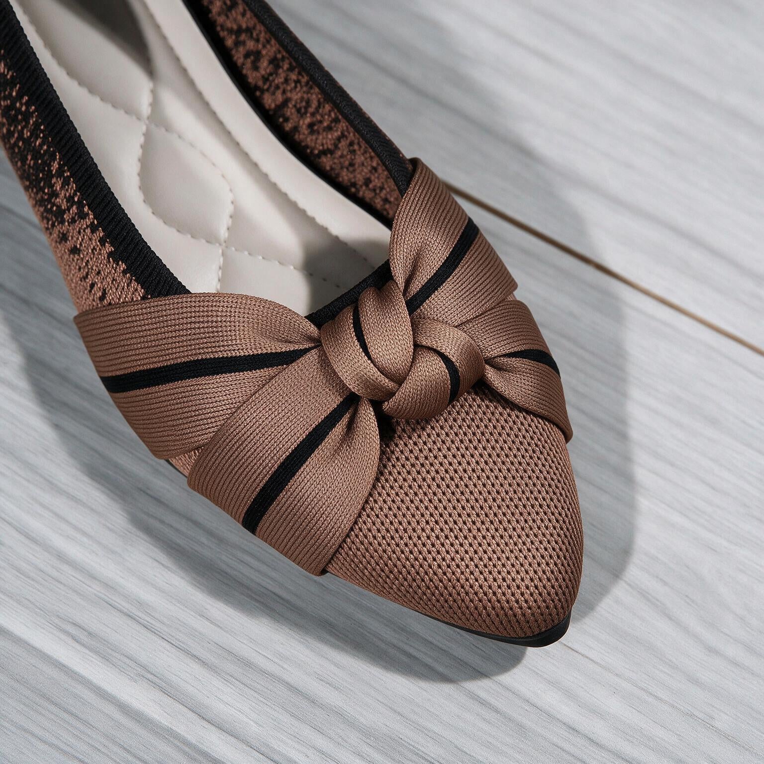 A close-up of a women's slip-on shoe with a pointed toe, featuring a large satin bow with black stripes on the vamp, a textured brown mesh toe box, and a quilted cream insole, set against a light gray wood floor.