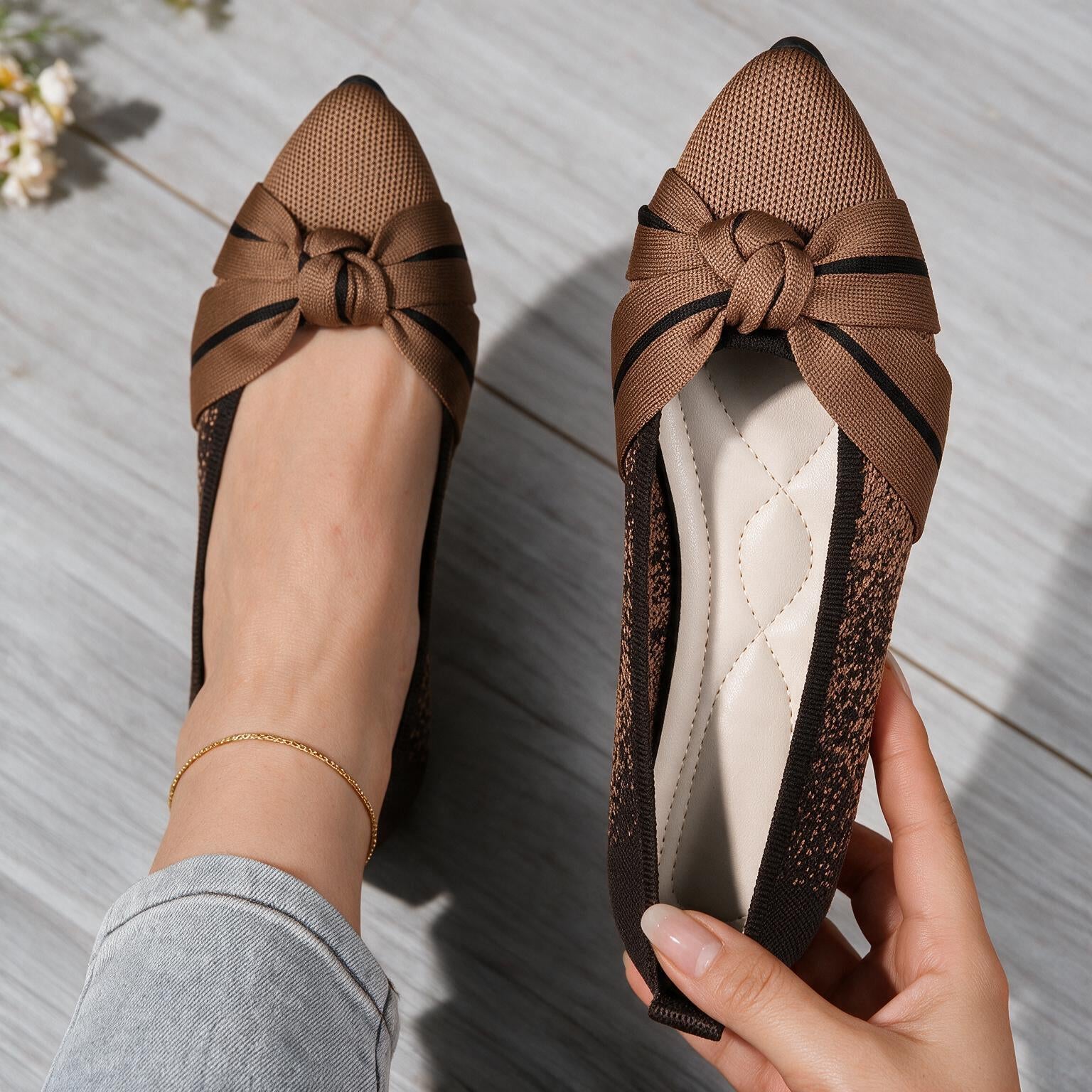 A pair of women's brown flat shoes with pointed toes and knotted bow details on the toes, shown on a person's foot and held in a hand over a gray wood floor.