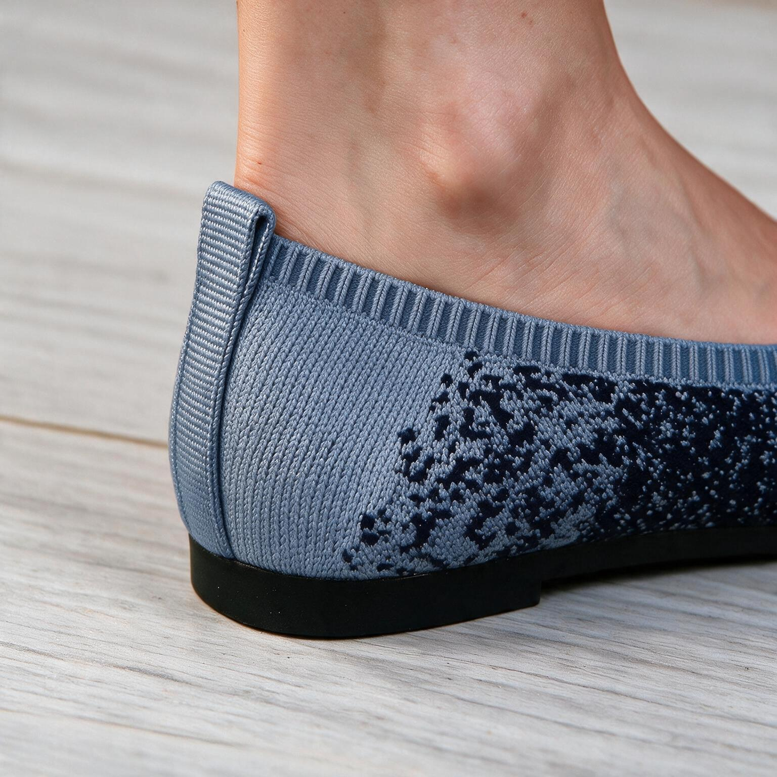 Close-up of a person's foot wearing a blue knit slip-on shoe with a black sole, standing on a light wood floor.